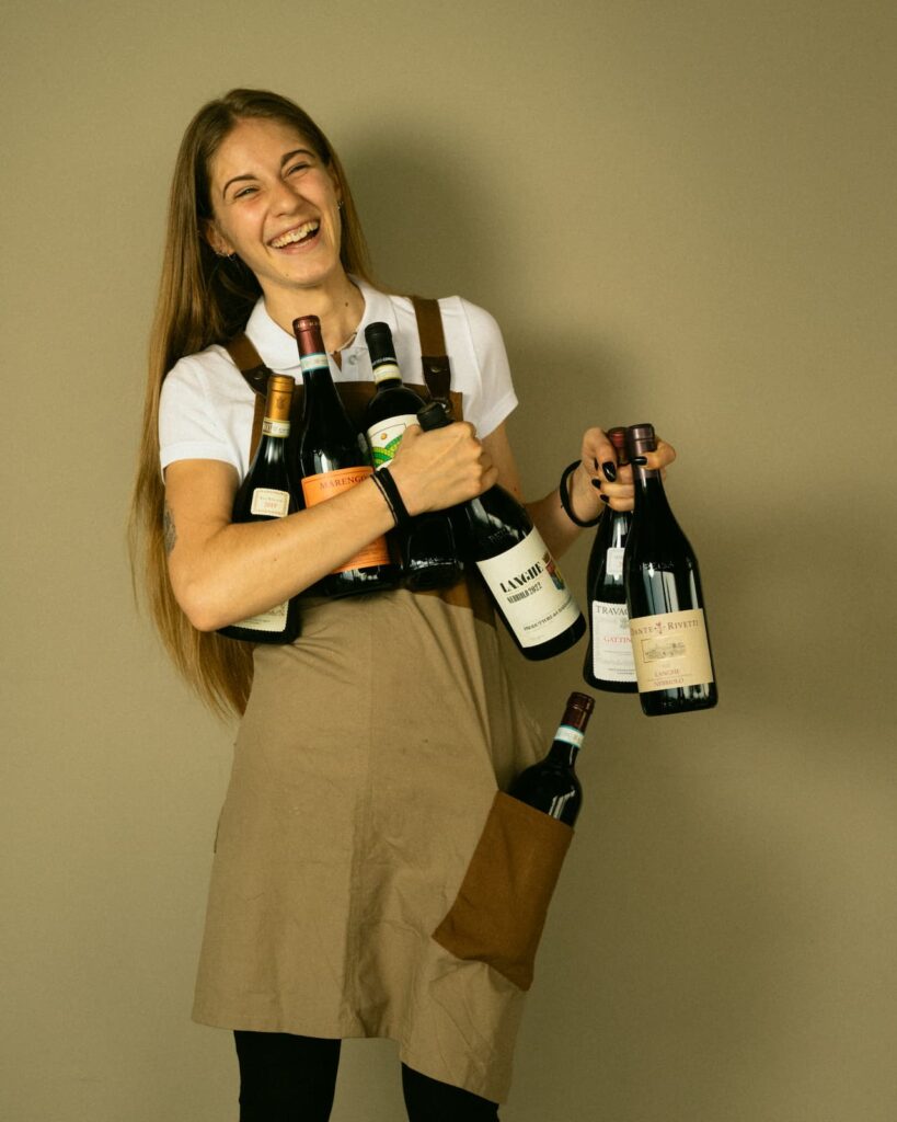 Giovane donna vestita da cameriera sorride reggendo in mano numerose bottiglie di vino